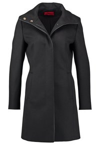 Manteau long noir avec col haut, deux poches latérales et boutons dorés sur l'épaule.