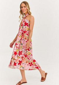 Žena kráčajúca v ružových kvetinových halter šatách s hnedými plochými sandálmi na pozadí bez vzoru.