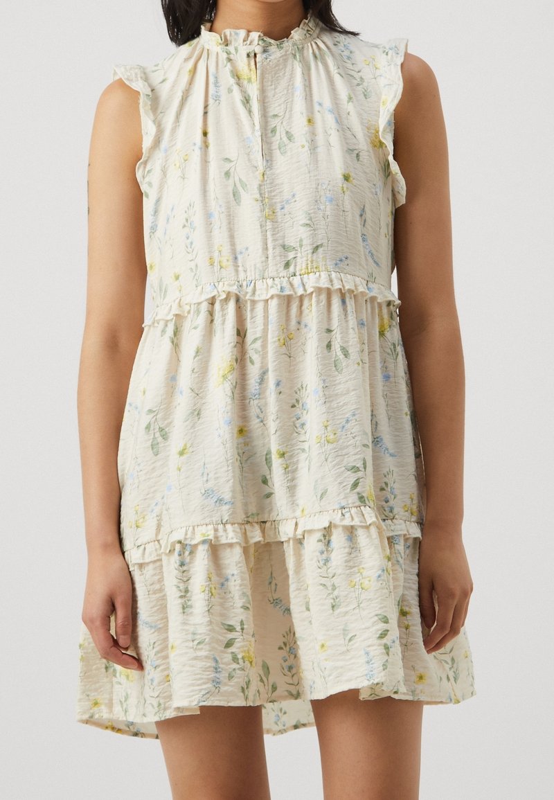 Robe crème sans manches avec bords à volants et petit imprimé floral en jaune pastel, bleu et vert, portée par une personne debout.