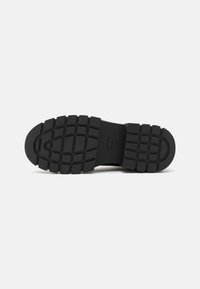 Suela de zapato de goma negra con un patrón texturizado de surcos profundos y un diseño ovalado en la parte delantera, que proporciona tracción y durabilidad.