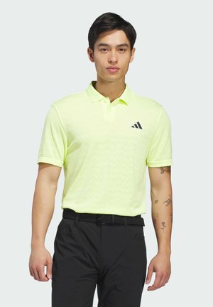 Homme portant un polo Adidas jaune clair et un pantalon noir, debout avec une expression neutre sur un fond uni.