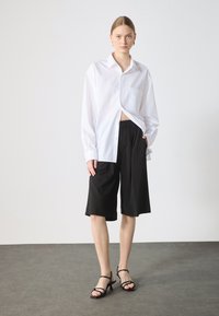 Camicia bianca oversize con bottoni, pantaloni corti neri su misura e tacchi neri con cinturini. La camicia ha una tasca sul petto e una vestibilità morbida.