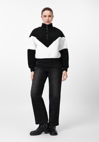 Mustvalge fliispullover, millel on poollukuga krae ja V-kujuline muster, kombineeritud mustade sirgelõikeliste teksade ja kontsaga pahkluu saapadega.