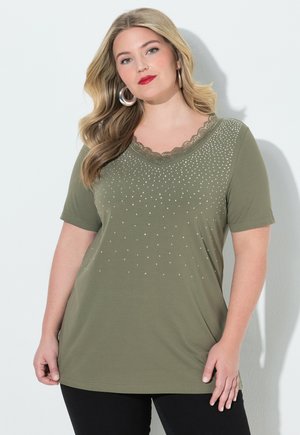 Kvinde med langt blondt hår iført store creoler og en olivengrøn top med korte ærmer, rhinsten og blondedetaljer.