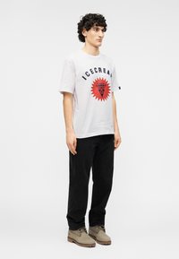 Λευκό βαμβακερό T-shirt με το κείμενο "ICECREAM" και γραφικά σε κόκκινο και μπλε. Συνδυασμένο με μαύρο παντελόνι και γκρι μποτάκια, σε όρθια στάση μπροστά σε απλό φόντο.