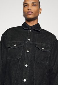 Giacca in denim nera con colletto, due tasche sul petto e bottoni a pressione argentati; tessuto strutturato con un aspetto leggermente usurato.