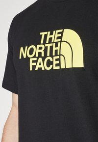 Černé bavlněné tričko s velkým žlutým nápisem "THE NORTH FACE," s výrazným logem na horní levé straně.