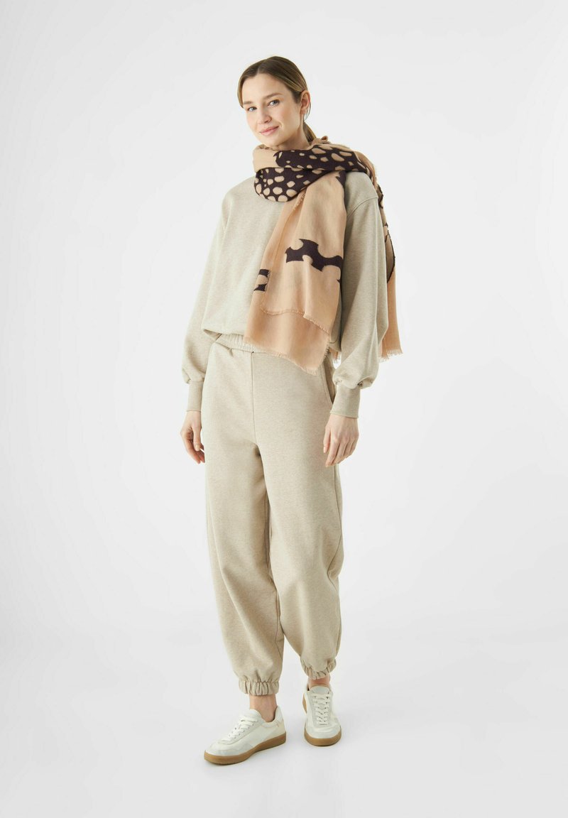 Sweatshirt en coton beige et joggers avec poignets élastiques, associés à une écharpe à motifs beige et marron. Baskets avec une partie supérieure blanche et une semelle marron.