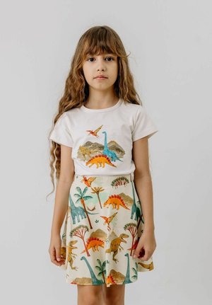 Mädchen mit langen braunen Haaren steht vor weißem Hintergrund, trägt ein weißes T-Shirt mit Dinosaurier-Aufdruck und einen passenden Rock mit Dinosaurier-Muster.