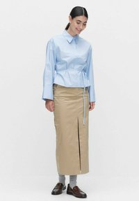Jasnoniebieska koszula z kołnierzykiem, z подсеченным w pasie i długimi rękawami, w zestawie z beżową spódnicą ołówkową z rozcięciem z przodu, noszona z szarymi skarpetkami i brązowymi mokasynami.