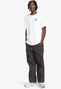 Camiseta de algodón blanca con texto azul y logo en el pecho, combinada con pantalones cargo negros de corte holgado y zapatillas deportivas grises.