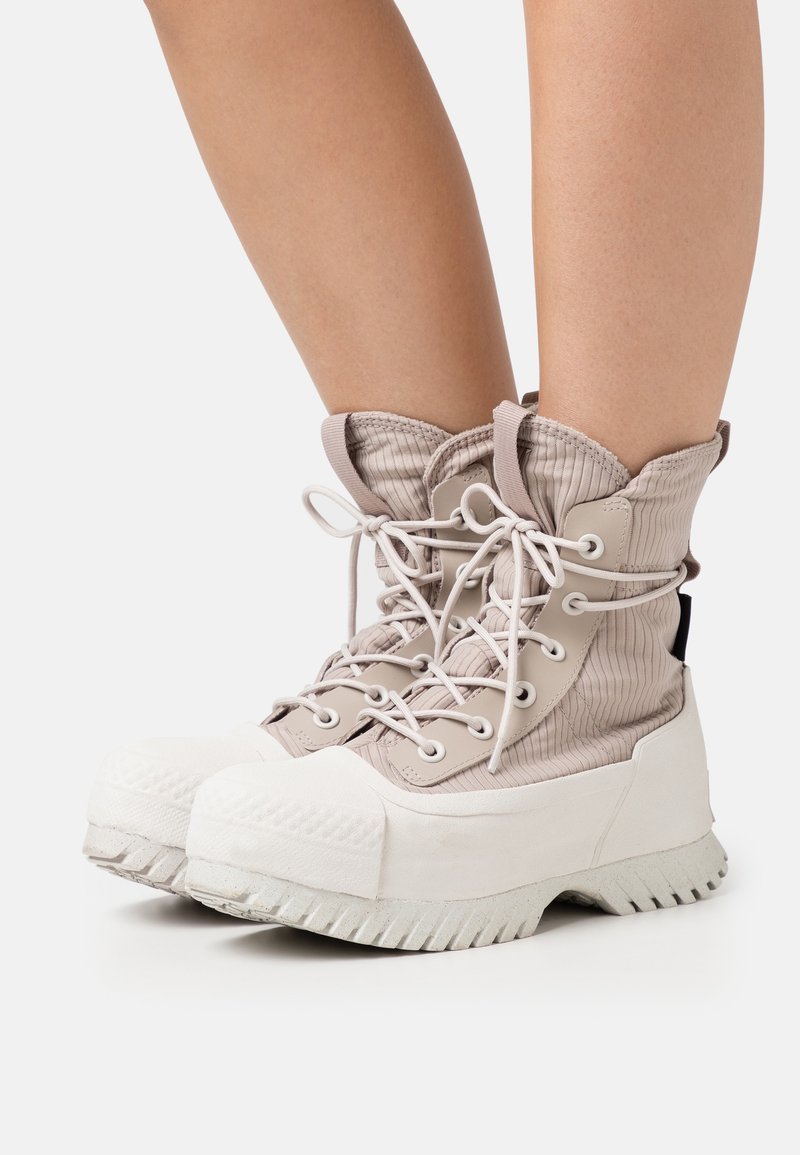 Bottes montantes beiges à lacets avec semelles épaisses blanches portées sur des jambes nues, montrant un tissu texturé et une semelle robuste pour une utilisation en extérieur.