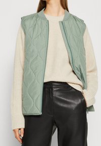 Jasnozielona pikowana kamizelka o fakturowym wykończeniu, z prążkowanym kołnierzem i zapięciem na zamek, połączona z kremowym swetrem z dzianiny i czarnymi skórzanymi spodniami.