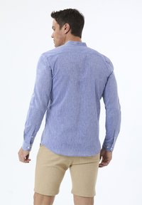 Camisa de manga larga azul claro con un acabado texturizado, detalles de botones en las mangas y un cuello relajado. Llevada con pantalones cortos beige.