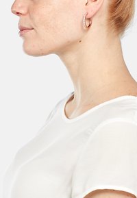 Ohrringe aus roségold mit klaren Kristallakzenten, die ein glattes, geschwungenes Design bieten. Trägt eine Person mit kurzen Haaren und einem weißen Hemd.