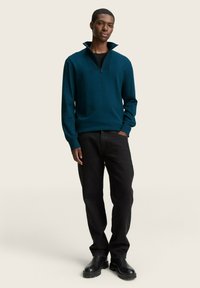 Türaus pullover mit Reißverschlusskragen, strukturierter Stoff, getragen über einem schwarzen Rundhalsausschnitt-Shirt, kombiniert mit geraden schwarzen Jeans und schwarzen Stiefeln.