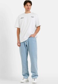 Camiseta blanca de manga corta con texto impreso en el pecho, combinada con jeans de corte holgado en azul claro y zapatillas blancas.