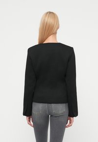 Μαύρο εφαρμοστό σακάκι με μακριά μανίκια, τετράγωνη λαιμόκοψη και δομημένη σιλουέτα. Φορεμένο πάνω από γκρι τζιν, παρουσιάζοντας ομαλή υφή υφάσματος.