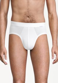 Uomo che indossa mutande di cotone bianche semplici, in piedi con le braccia rilassate contro uno sfondo chiaro e uniforme.
