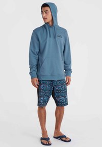 Capuchontrui in teal met een voorzak en trekkingkoord; gecombineerd met meerkleurige gestreepte shorts en slippers.