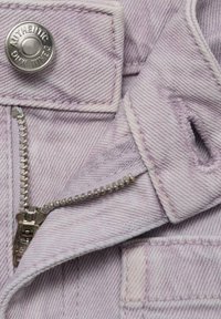 Giacca in denim di un leggero color viola con zip silver, bottoni a pressione in metallo e un tessuto leggermente strutturato. Include una tasca e dettagli di cucitura.