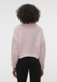 Personne aux cheveux bouclés vue de dos, portant un pull en tricot texturé rose et un pantalon noir sur un fond blanc uni.