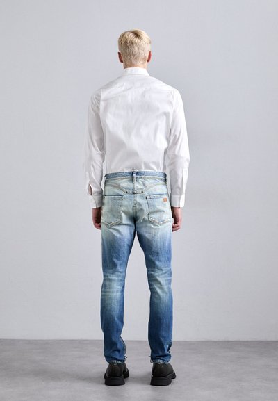Persona bionda di spalle, con una camicia bianca a maniche lunghe, jeans chiari sbiaditi e scarpe nere, che si trova in piedi contro un muro grigio semplice.