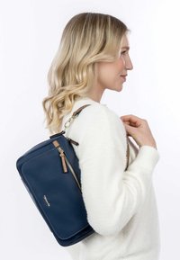 Navyblaue Leder-Rucksack mit glatter Textur, goldfarbigem Reißverschluss und braunem Akzentgurt, der an einer Schulter getragen wird. Rechteckige Form.