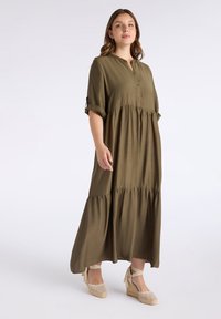 Langes olivengrünes Kleid aus weichem Stoff mit einer Knopfleiste vorne, kurzen Umschlagsärmeln und einem gestuften, ausgestellten Saum. Kombiniert mit Espadrilles-Wedges.