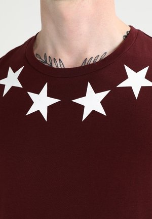Gros plan sur le cou d'une personne avec des tatouages de feuilles, portant une chemise bordeaux ornée de cinq étoiles blanches le long de l'encolure.