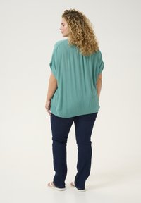 Blouse teal ample avec un tissu texturé, manches courtes roulées et un ourlet arrondi. Associée à un jean bleu foncé et des sandales claires.