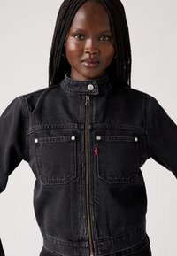 Chaqueta de mezclilla negra con cuello, cremallera frontal y dos bolsillos en el pecho. Presenta botones a presión blancos y un sutil detalle del logo de Levi's.