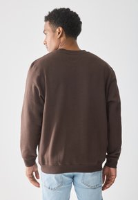 Man die zich omkeert en een brown sweatshirt en lichte blauwe jeans draagt tegen een effen witte achtergrond.