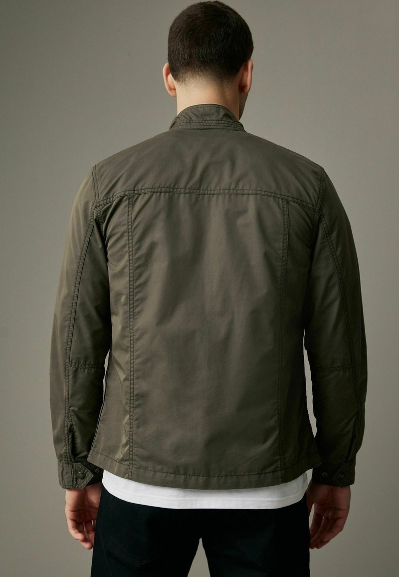 Veste vert olive avec un col, des poignets boutonnés et une coupe droite. Fabriquée en tissu léger avec une texture lisse et des coutures visibles.