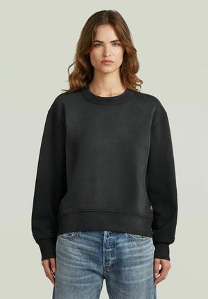 Donna con capelli castani di media lunghezza, che indossa una felpa nera semplice e jeans blu, in piedi davanti a uno sfondo grigio chiaro.