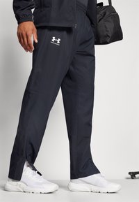 Mann in schwarzen Under Armour Jogginghosen, weißen Sneakers, hält eine schwarze Sporttasche an seiner Seite vor einem schlichten Hintergrund.