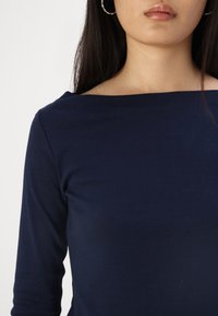 Tamsiai mėlynas ilgas rankovės topas su plačiu iškirpimu ir lygiomis, minkštomis tekstūromis, turintis minimalų siuvimą ir be matomų raštų.