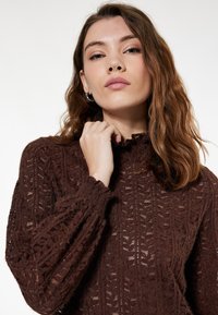 Ruskea neulevillapaita, jossa on teksturoitu lehtikuosi, korkea kaulus ja rypytetyt hihat. Valmistettu pehmeästä ja kevyestä materiaalista.