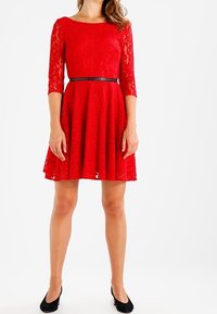 Robe en dentelle rouge avec des manches trois-quarts, jupe évasée, col rond et ceinture noire avec des accents argentés. Portée avec des chaussures noires.