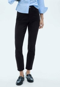 Črne kavbojke iz denima s ozkim krojem, standardnim pasom in petimi žepi, v kombinaciji s črnimi mokasini in svetlobluzo srajco na gumbe.