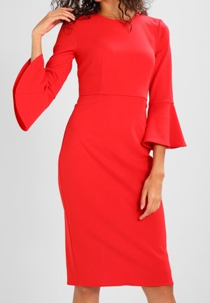 Femme portant une robe rouge vif cintrée à la longueur du genou avec des manches évasées trois-quarts, debout devant un fond gris clair.
