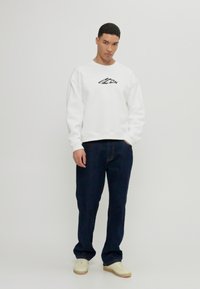 Sweatshirt blanc avec un logo noir, associé à un jean bleu foncé et des baskets beiges. Design simple, coupe décontractée et tissu texturé.