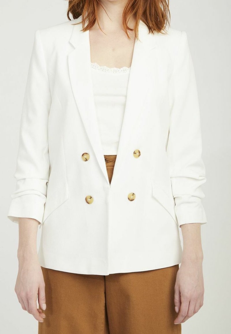 Blazer blanc croisé en tissu texturé, avec des boutons dorés et des revers crantés, associé à un pantalon marron.