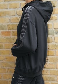 Černá mikina s kapucí a dlouhými rukávy, svislé bílé proužky na obou pažích a žebrované manžety. Měkká látka a volný střih.