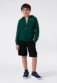 Camisola com capuz verde com fecho zip e bolsos laterais, logótipo branco no peito e na manga, combinada com calções pretos e ténis com detalhes azuis.