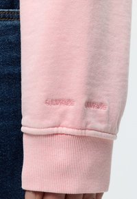 Κοντινό ενός μανικιού ροζ φούτερ με ριμπ μανσέτα και διακριτική κεντητική κοντά στον καρπό, φορεμένο με σκούρο μπλε τζιν.