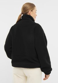 Čierny fleecový pulover s vysokým golierom, spadnutými ramenami a voľným strihom. Mäkká textúra bez viditeľných vzorov alebo hardvérových detailov.