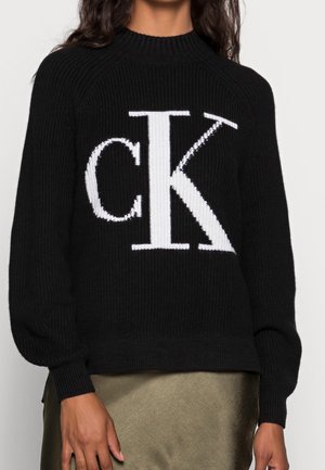 Vrouw die een zwarte gebreide trui draagt met grote witte "cK" logo letters op de voorkant en een metallic olijfgroene rok.