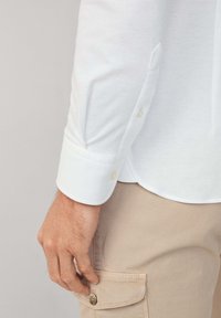 Biała koszula z długim rękawem i zapięciem na guziki przy mankietach, zestawiona z beżowymi spodniami cargo. Koszula ma gładką teksturę i zaokrąglony dół.