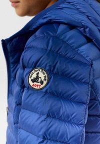 Blauwe pufferjack met doorgestikt ontwerp, capuchon en een ronde patch met tekst en een grafisch logo op de linker schouder.